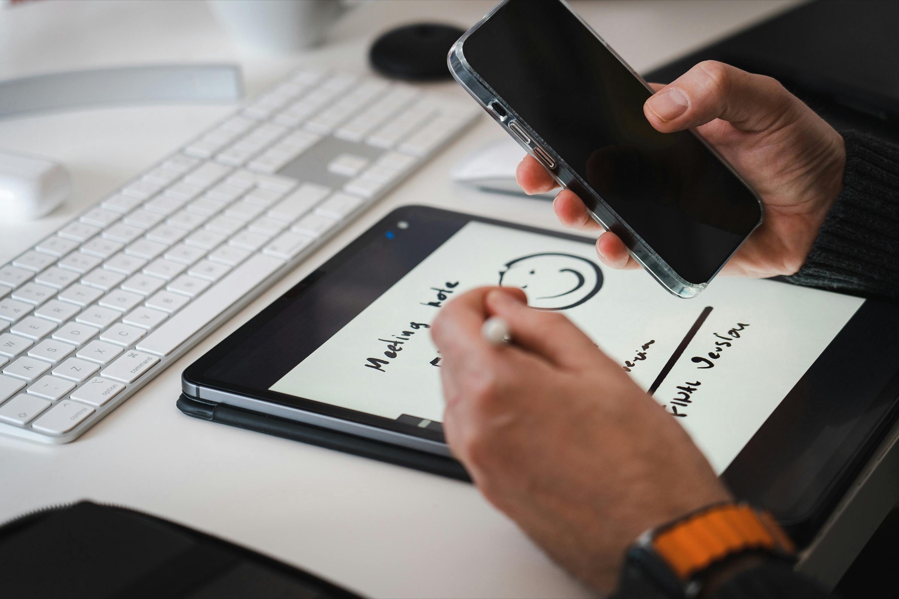 Hands using a stylus on a tablet while holding a phone above a planning workspace.